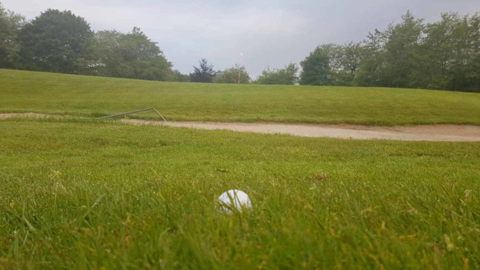 Im Vordergrund ein weißer Golfball in etwas höherem Gras, dahinter der Bunker. Etwas erhöht im Hintergrund ist die Fahne zu erkennen, das Loch, in dem sie steckt, sieht man nicht.