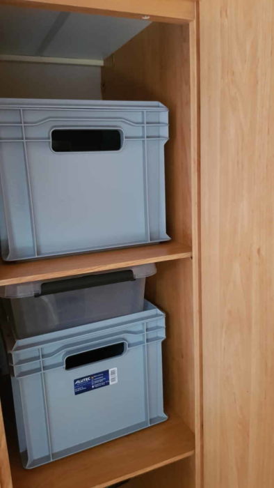 In einem schmalen Schrank mit Ahornfurnier stehen zwei graue Kunststoffkisten in je einem Fach. Auf der unteren Kiste findet noch eine flache, halbdurchsichtige Kunststoffkiste Platz.