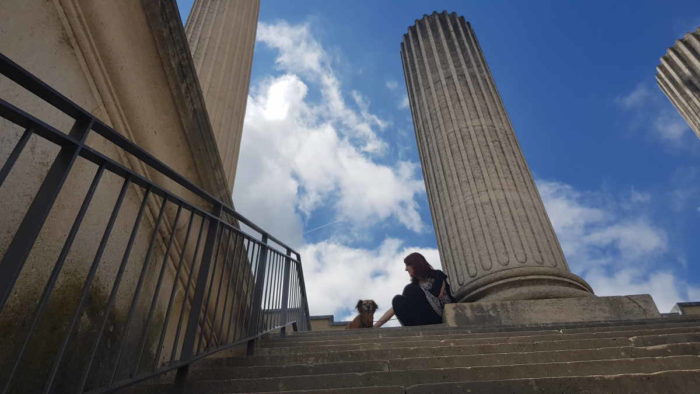 Aus der Froschperspektive aufgenommen: Gesche und Lexi sitzen am oberen Ende der Treppe des Hafentempels im archäologischen Park Xanten. Dorische Säulen ragen in den leicht bewölkten Himmel.