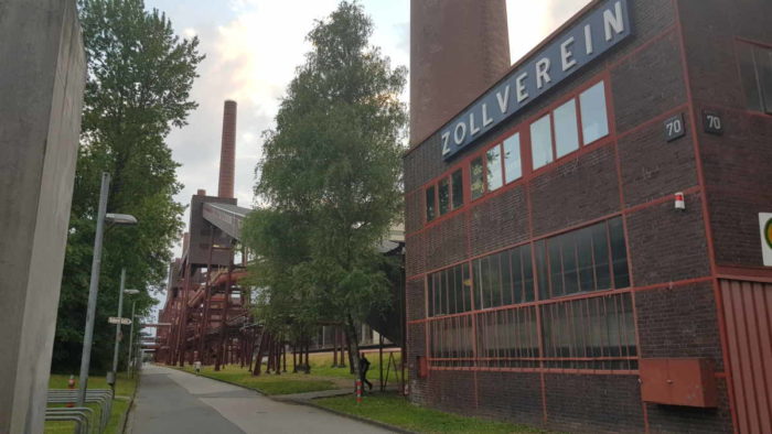 Ein Gebäude der Zeche Zollverein, im Hintergrund mehrere Bäume und technische Anlagen, die heute nicht mehr in Betrieb sind.