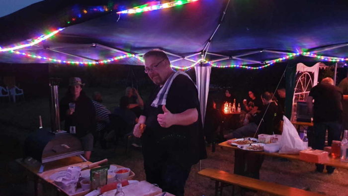 Sven steht unter dem mit Lichterketten beleuchteten Pavillon und macht die Daumen hoch-Geste. Neben ihm stehen Utensilien und Zutaten für Pizza. (Foto von Rüdiger)
