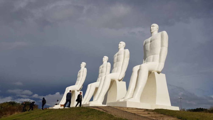 Die Skulptur "Der Mensch und das Meer" am Strand von Esbjerg: Vier acht Meter große Figuren aus weißem Beton. Sie sehen männlich aus und sind sitzend dargestellt. Die Sonne scheint auf die Skultpuren, im Hintergrund dunkle Wolken.