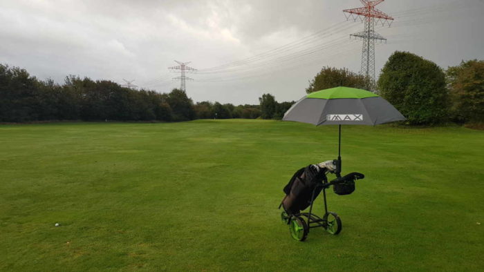 Mein Golftrolley steht neben dem Ball auf der Bahn 5 des Husumer Golfclubs. Am Trolley ist ein grau-grüner Regenschirm befestigt, der Himmel ist grau. Man kann es nicht erkennen, aber es regnet.