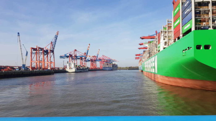 Blick in den Containerhafen von Hamburg, rechts ist ein großes, grünes Containerschiff im Anschnitt zu sehen. Am linken Bildrand Containerbrücken. Es ist sommerliches Wetter mit viel Sonnenschein und blauem Himmel.
