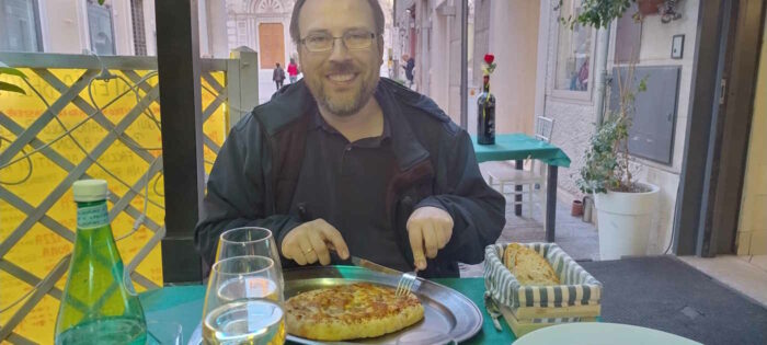 Portrait von mir, wie ich meine Pizza anschneide. Auf dem Tisch stehen außerdem eine Flasche Wasser und zwei Gläser mit Weißwein sowie ein Korb mit etwas Brot.