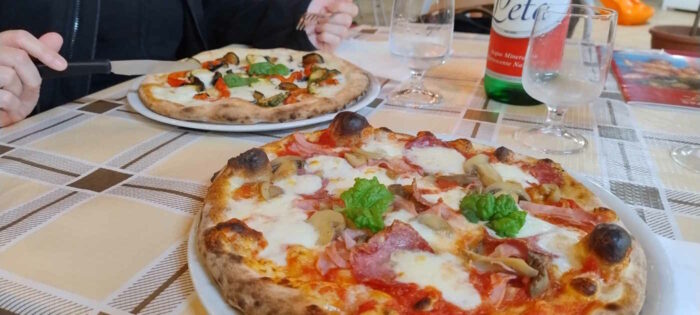 Nahaufnahme einer Pizza Capricciosa. Dahinter ist Gesches vegetarische Pizza zu sehen. 