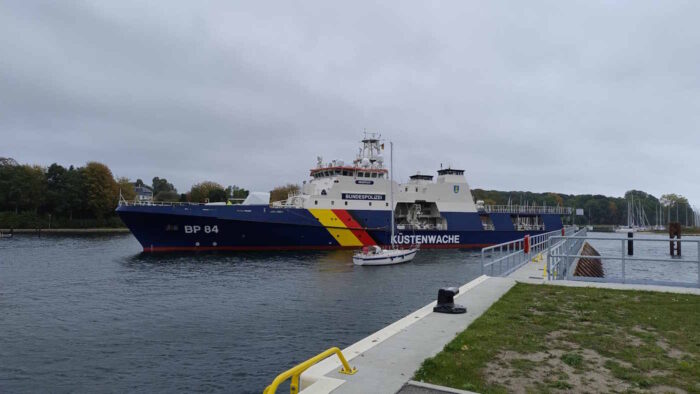 In der Bildmitte ein Schiff der "Potsdam"-Klasse. Der Rumpf ist blau lackiert und trägt sowohl die Kennung "BP 84" als auch den Schriftzug "Küstenwache". Die Decksaufbauten sind weiß. Vor dem Schiff liegt ein etwa 10 Meter langes Segelboot im Wasser. Es wirkt winzig.