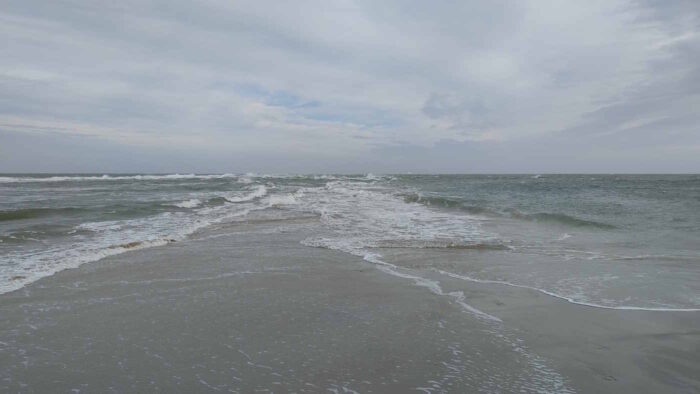 Die Sandbank von Skagen, an der sich Nord- und Ostsee treffen. Von rechts und links überspülen schäumende Wellen die Sandbank, der Himmel ist grau.