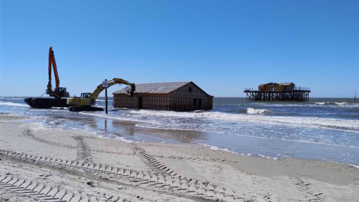 Rechts im Hintergrund steht der Rest eines St. Peter-Ordinger Pfahlbaus in der Nordsee. In der Bildmitte schwimmt der andere Teil gesichert von zwei Baggern an der Wasserkante. 