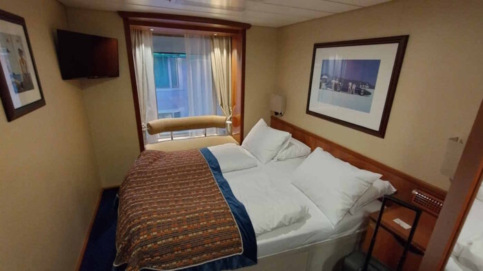 Eine Innenkabine der Color Magic. Der Raum wird von einem Doppelbett dominiert. Darüber hängt ein Bild. Der Nachttisch ist in dunklem Braun gehalten. Im Bildhintergrund ist ein Fenster mit einer gepolsterten Bank zu sehen. Durch die Gardine kann man die gegenüberliegende Kabine erkennen. In der linken oberen Ecke des Bildes hängt ein Fernseher.