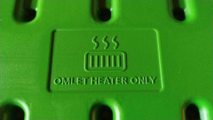 Ein grünes Plastikteil unseres Hühnerstalls in Nah-Aufnahme. Zu sehen ist ein Piktogram, das eine Heizung darstellt, und der Schriftzug