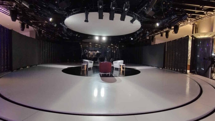 Ein kreisrundes Fernsehset mit weißen Boden. Darin erkennbar sind die Schienen, um LED-Wände zu positionieren. In der Mitte steht eine kleine Sitzgruppe, an der Decke hängen etliche Scheinwerfer. 
