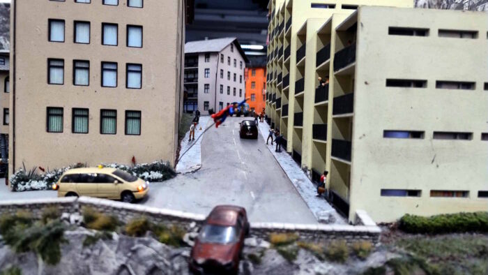 Straßenszene im Miniaturwunderland: Zwischen Häusern stehen kleine Figuren, ihre Körperhaltung drückt Erstaunen oder Entsetzen aus. Denn im Bildvordergrund sieht es so aus, als bräche gerade ein Auto durch eine Leitplanke und stürze die Klippe runter. In der Bildmitte und falsch fokussiert "fliegt" aber schon Superman zur Rettung.