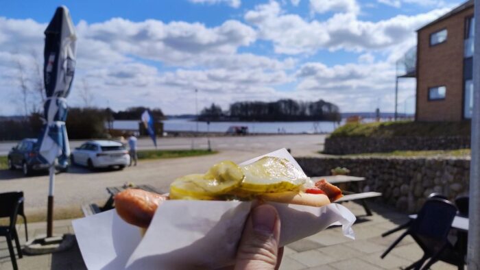 Im Vordergrund ist ein dänischer Hotdog mit Gurken drauf zu sehen, der in einer kleinen Papiertüte steckt und von einer Hand vor die Kamera gehalten wird. Dahinter erkennt man unscharf den Parkplatz, ein Haus und die Ochseninseln. Die Sonne scheint und am Himmel sind einige kleine Wolken.