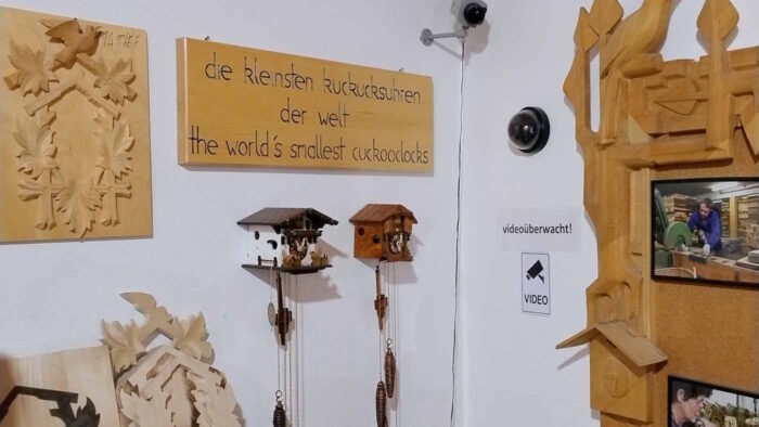 Eine Ecke in einem Museum. Zwei sehr kleine Kuckucksuhren hängen an einer Wand zwischen anderen Exponaten und zwei Überwachungskameras. Ein Schild weist darauf hin, dass es sich um die kleinsten Kuckucksuhren der Welt handelt.
