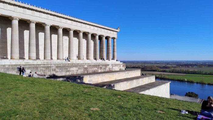 Die Walhalla bei Regensburg: Eine Gedenkhalle im Stil eines altgriechischen Tempels. Trotz Absperrungsbändern sitzen Leute auf den Stufen oder auf der Grünanlage. Die Sonne scheint, der Himmel ist blau. Die Aussicht auf das Donautal ist sehr beeindruckend.