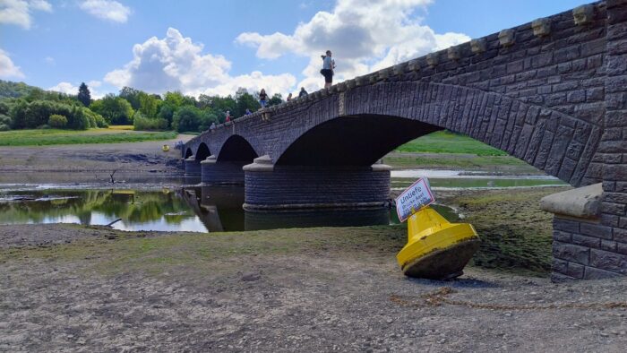 Die Brücke von Alt-Asel taucht immer bei niedrigem Wasserstand des Edersees auf. Das Bild zeigt die historische Brücke und davor eine gelbe Boje im Sand. An der Boje ist ein Schld angebracht, dass vor einer Untiefe durch die Brücke warnt.